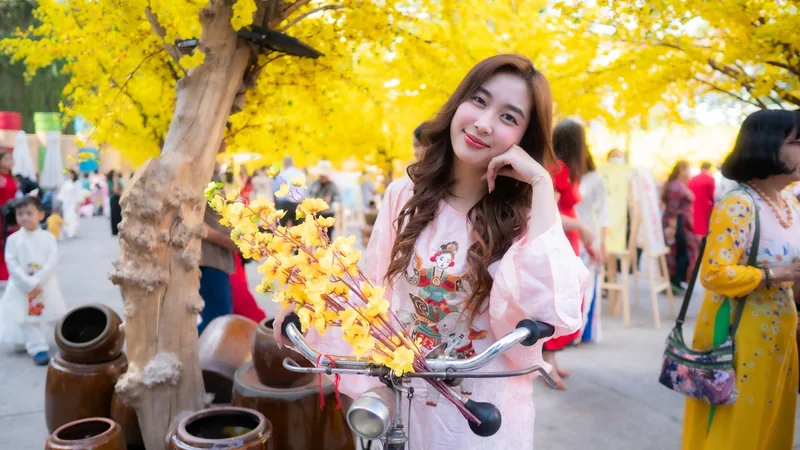 Một cô gái trẻ mặc trang phục truyền thống màu hồng tạo dáng bên xe đạp dưới hoa mai vàng trong lễ hội Tết ở Việt Nam.